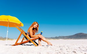 Beautiful young woman apllying sunblock to her legs while sitting on a beach in summer.